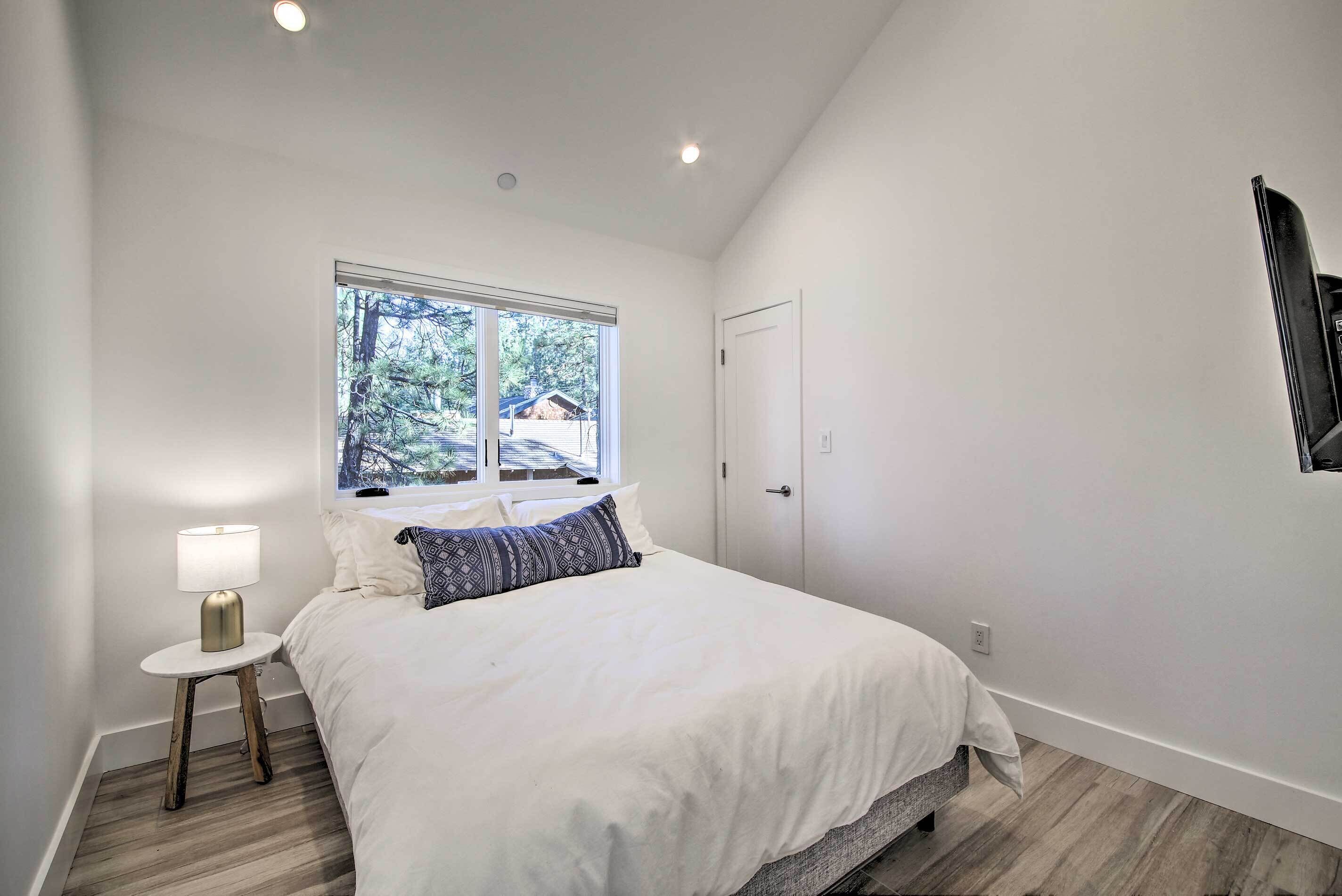Une chambre à coucher avec un grand lit, une table de chevet avec une lampe, une fenêtre donnant sur des arbres et une porte.