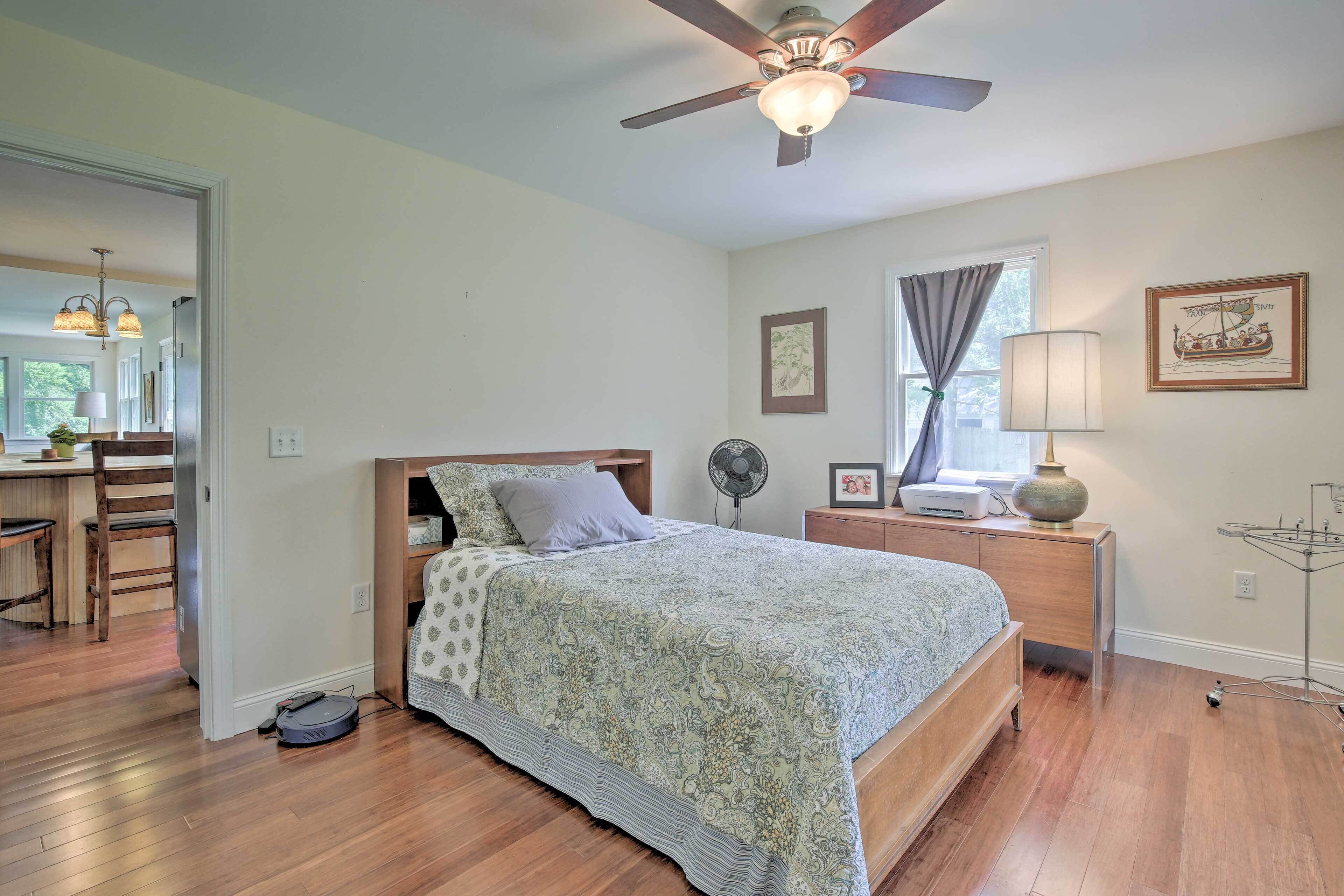 A bedroom with a bed, a ceiling fan, a desk with a chair, a window with curtains, and a wall-mounted lamp.