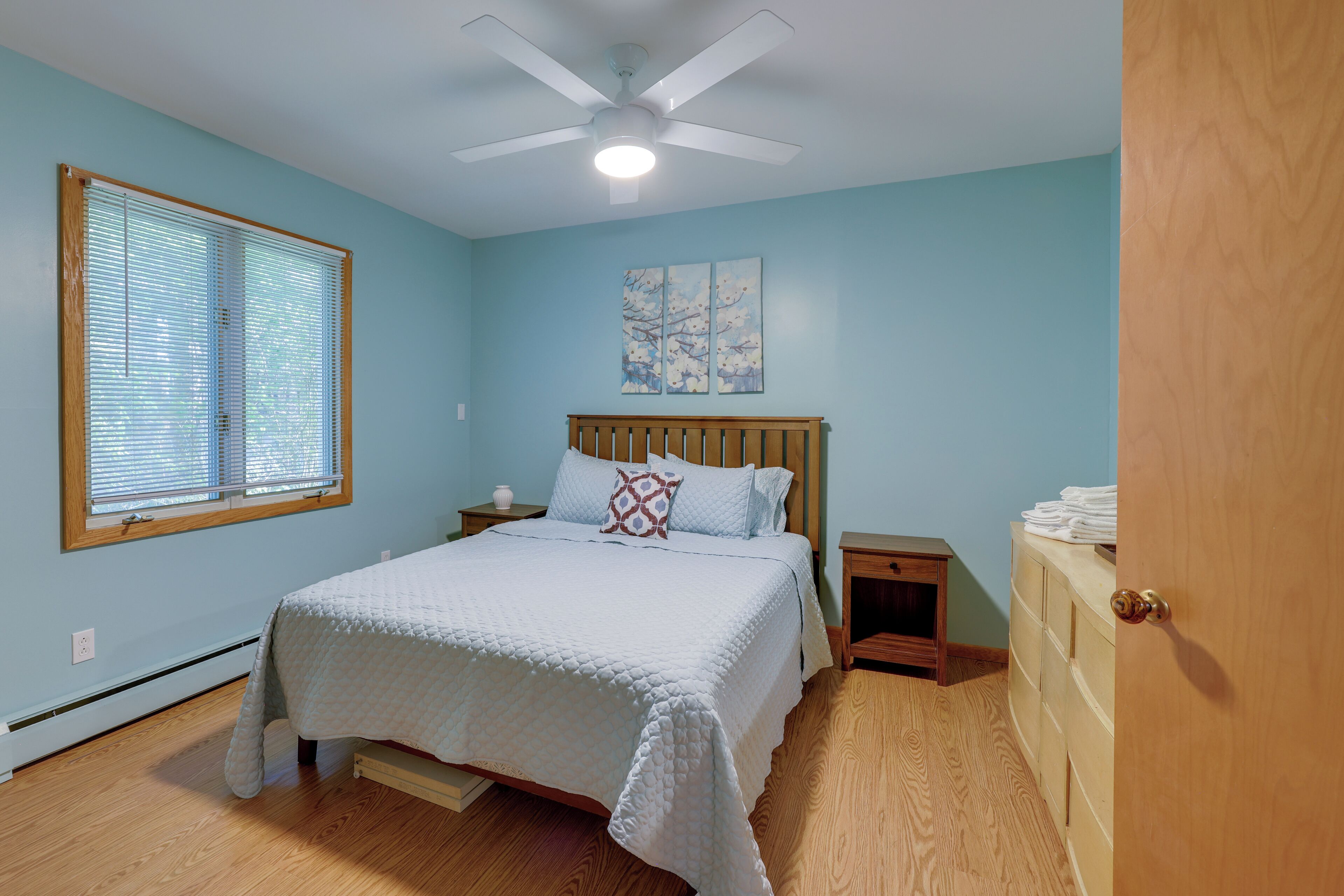 Une chambre à coucher comprenant un lit, une table de chevet, une tête de lit en bois, un ventilateur de plafond, une fenêtre avec des stores et une porte.