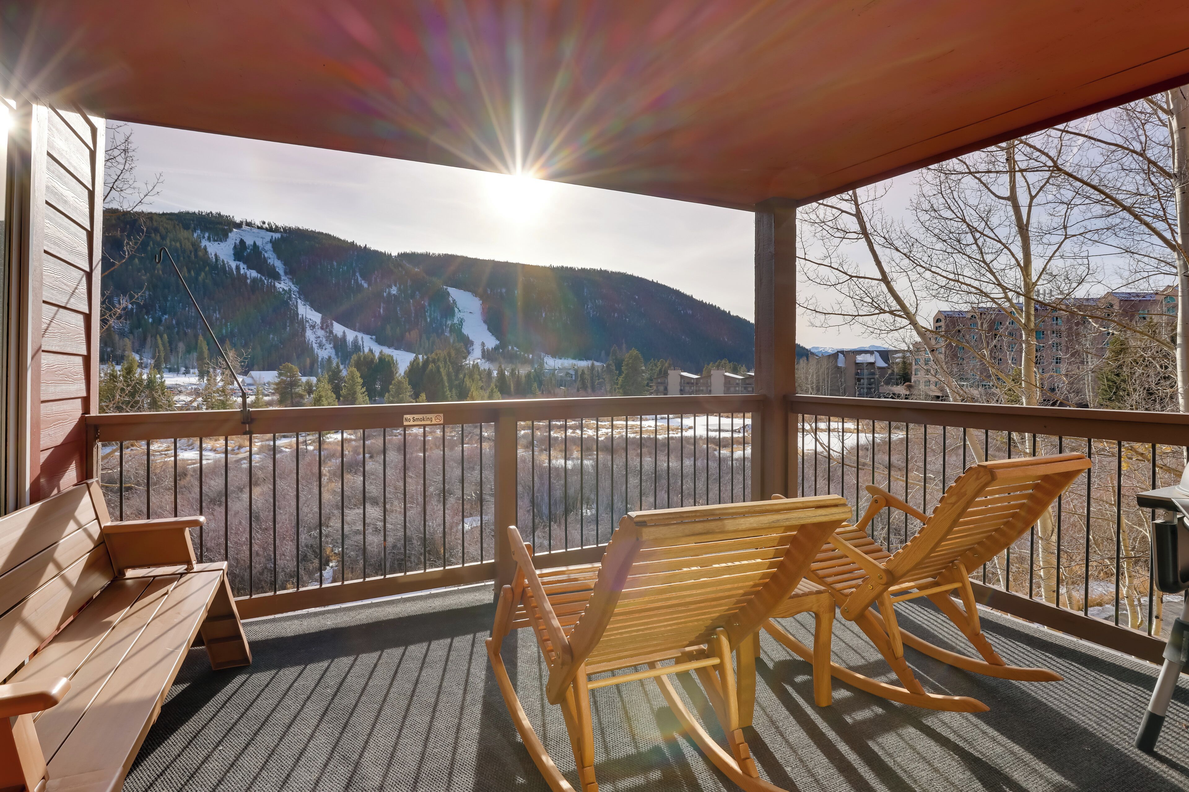 A wooden deck with a bench and rocking chairs, overlooking a snowy mountain and a parking lot.
