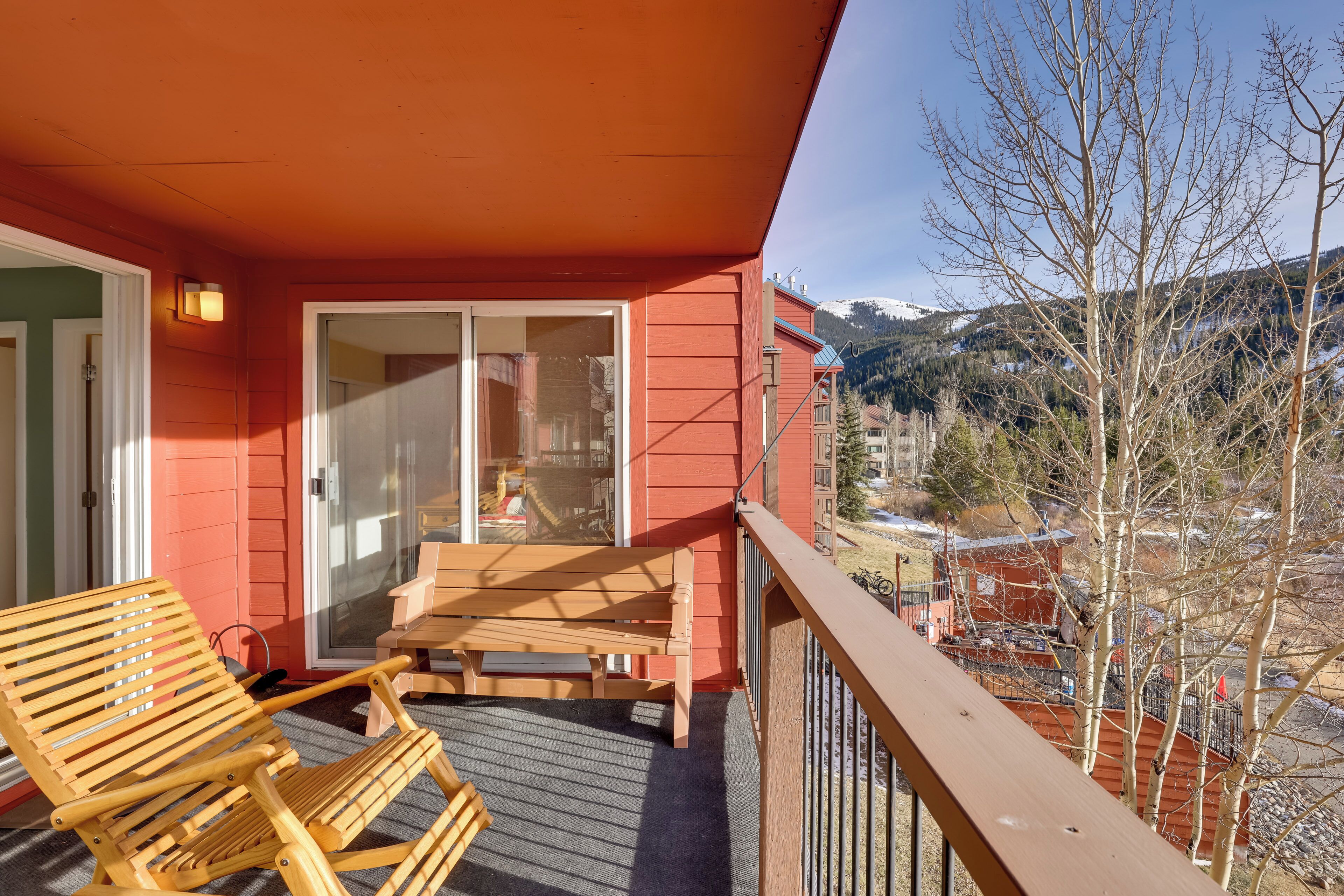 A balcony with a wooden chair and table, a red exterior wall, and a view of a snowy mountain and trees.
