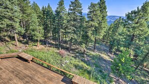 A wooden deck with a bench, surrounded by a dense forest and a clear view of distant mountains. - Spacious Evergreen Home w/ Picturesque Views (Evergreen)