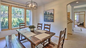 A dining area with a wooden table, four chairs, a bench, and a chandelier. Large windows offer a view of trees and a field. A painting is on the wall. - Spacious Evergreen Home w/ Picturesque Views (Evergreen)
