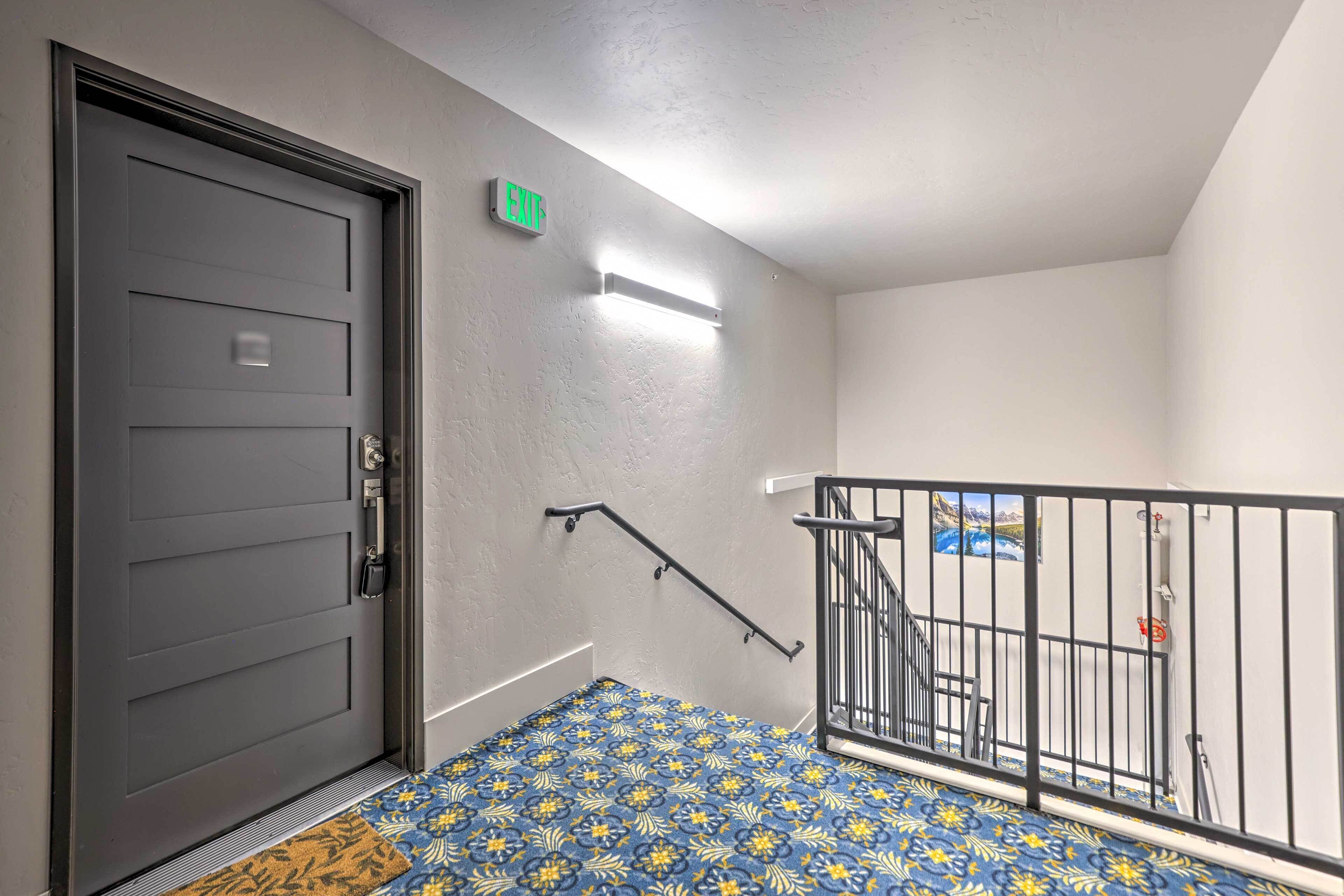 A modern apartment entrance with a dark gray door, an exit sign, a staircase with a metal railing, and a patterned blue and yellow floor mat.