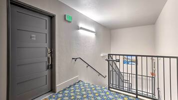 A modern apartment entrance with a dark gray door, an exit sign, a staircase with a metal railing, and a patterned blue and yellow floor mat.