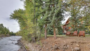 Un chalet au bord de la rivière, doté d’une terrasse en bois et de fauteuils Adirondack, entouré d’arbres et bordé d’une rive rocheuse.