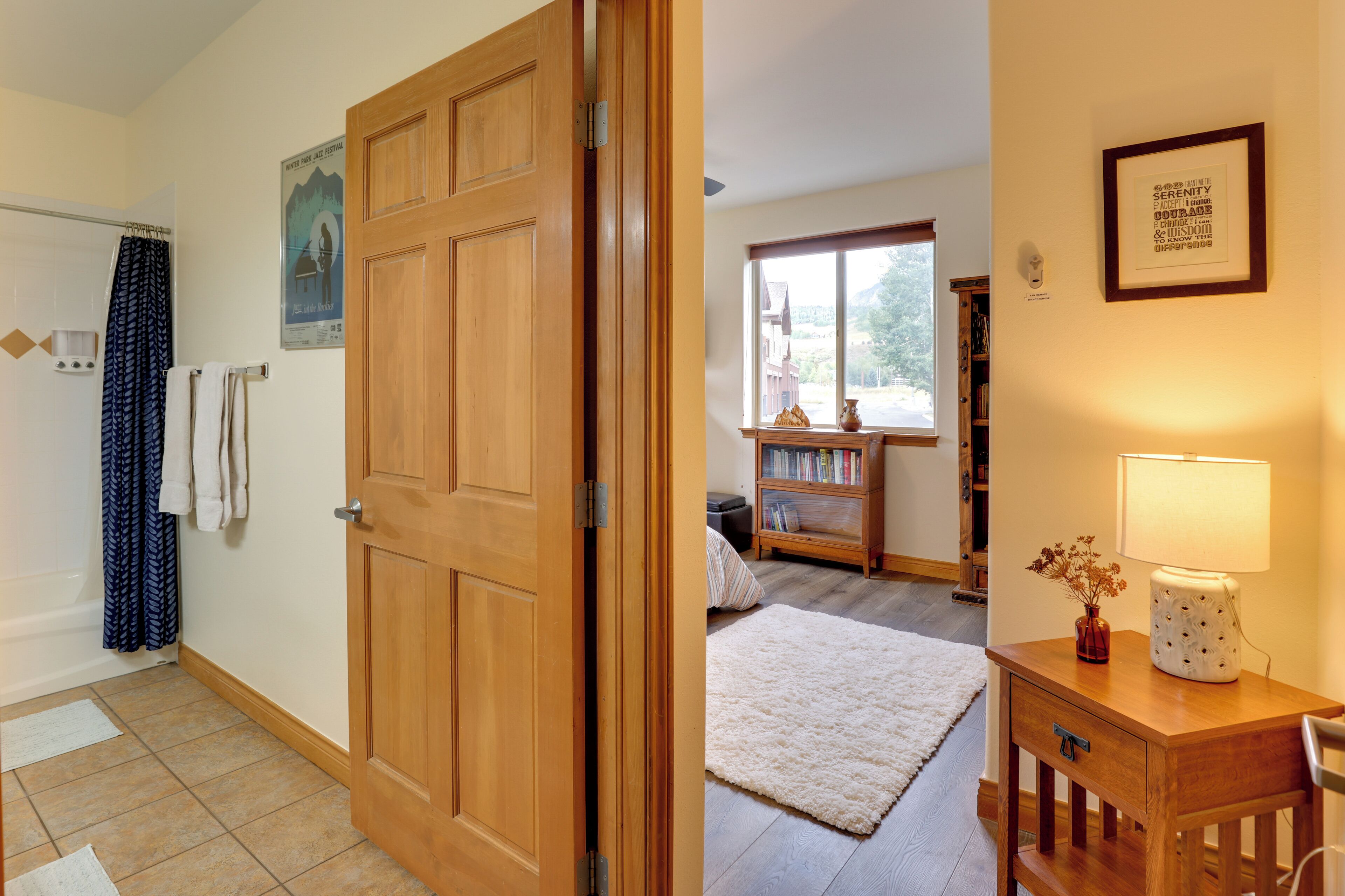 Una habitación con puerta de madera, alfombra blanca, una mesita de noche de madera con lámpara y una estantería.