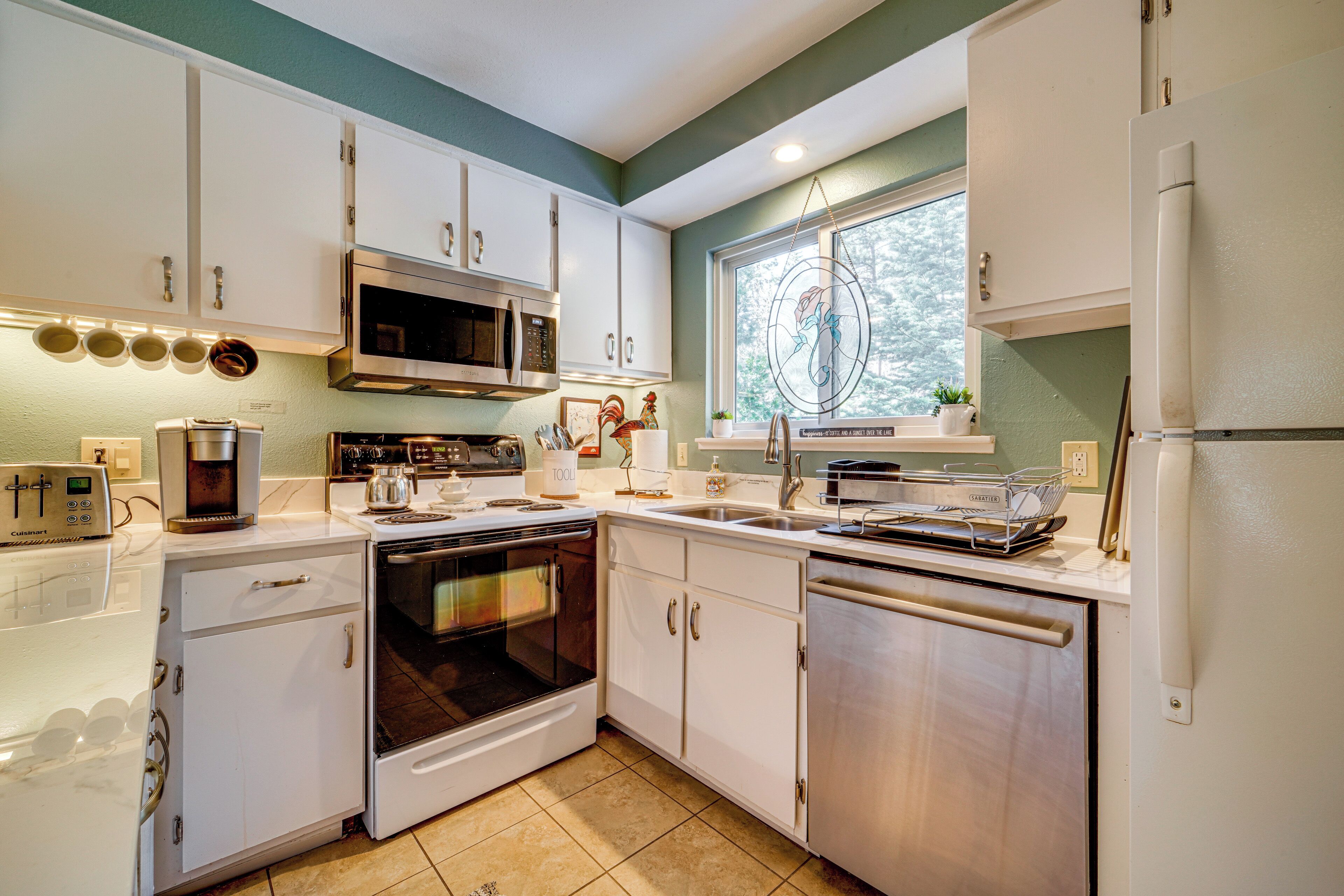 A modern kitchen with white cabinets, stainless steel appliances, and a window with a view of trees.