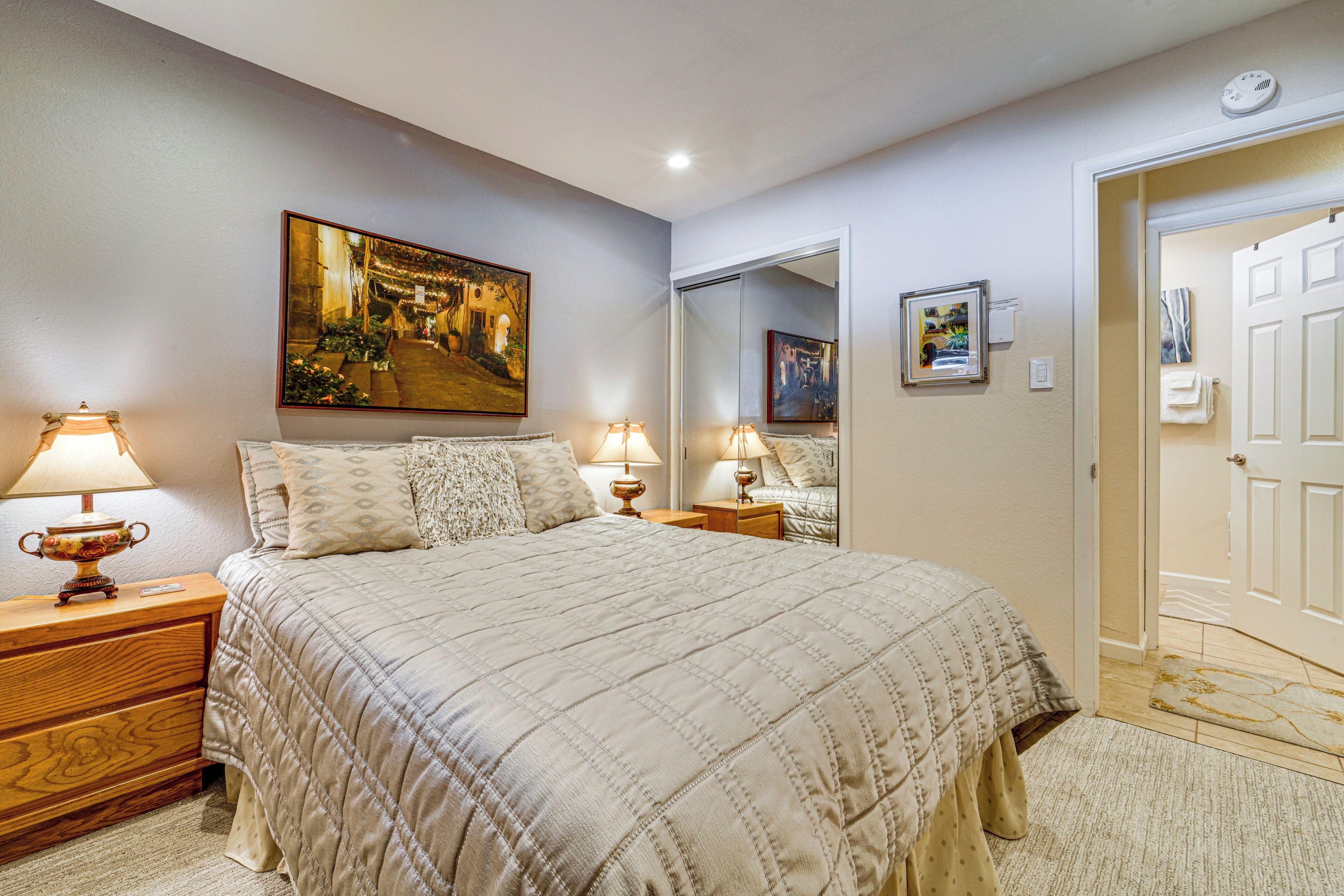 Une chambre à coucher avec un grand lit, deux tables de chevet avec des lampes, un tableau accroché au mur, un miroir et une porte donnant sur une autre pièce.
