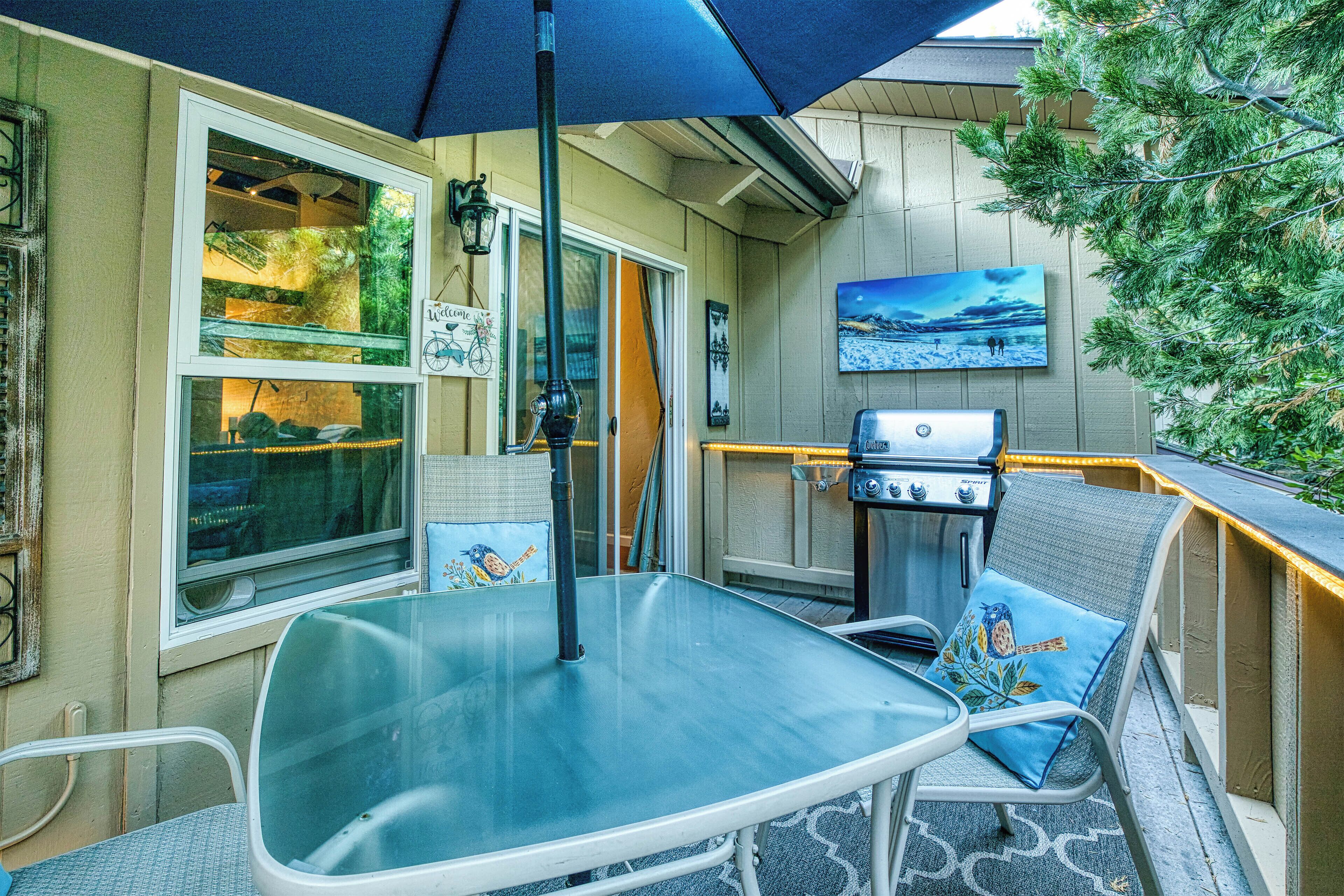 An outdoor patio area with a glass table, metal chairs, a blue umbrella, a grill, and a painting on the wall.
