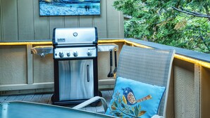 A barbecue grill and a chair with a bird design, both on an outdoor deck with a wooden wall and a painting above the grill. - Artists Tahoe Retreat: Pool, Art & Ski Access! (Incline Village)