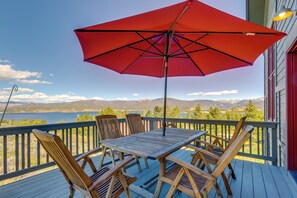 A wooden deck with a table and chairs, a red umbrella, and a view of mountains and a lake. - Granby Home: Stunning Mountain & Lake Views! (Granby)