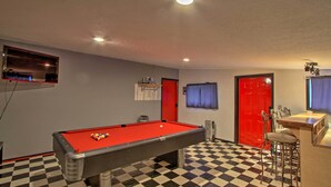 Ein Billardtisch mit roter Spielvliesdecke, ein Schachbrettmuster-Boden und eine Bar mit Stehtischen.