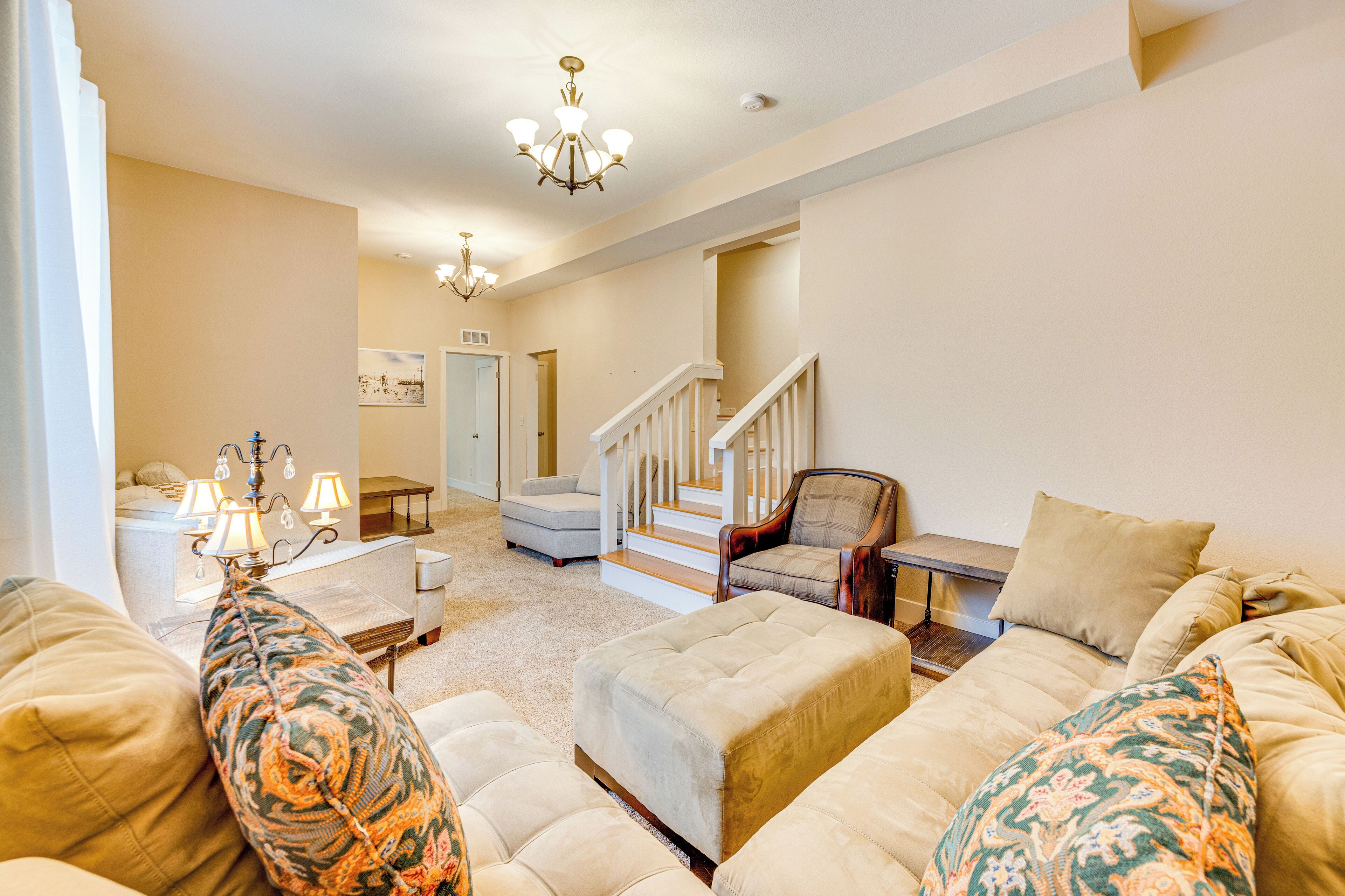A spacious living room with a large beige sofa, a patterned armchair, and a wooden coffee table. A staircase leads to an upper level, and a chandelier hangs from the ceiling.
