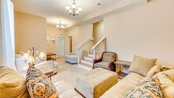 A spacious living room with a large beige sofa, a patterned armchair, and a wooden coffee table. A staircase leads to an upper level, and a chandelier hangs from the ceiling.