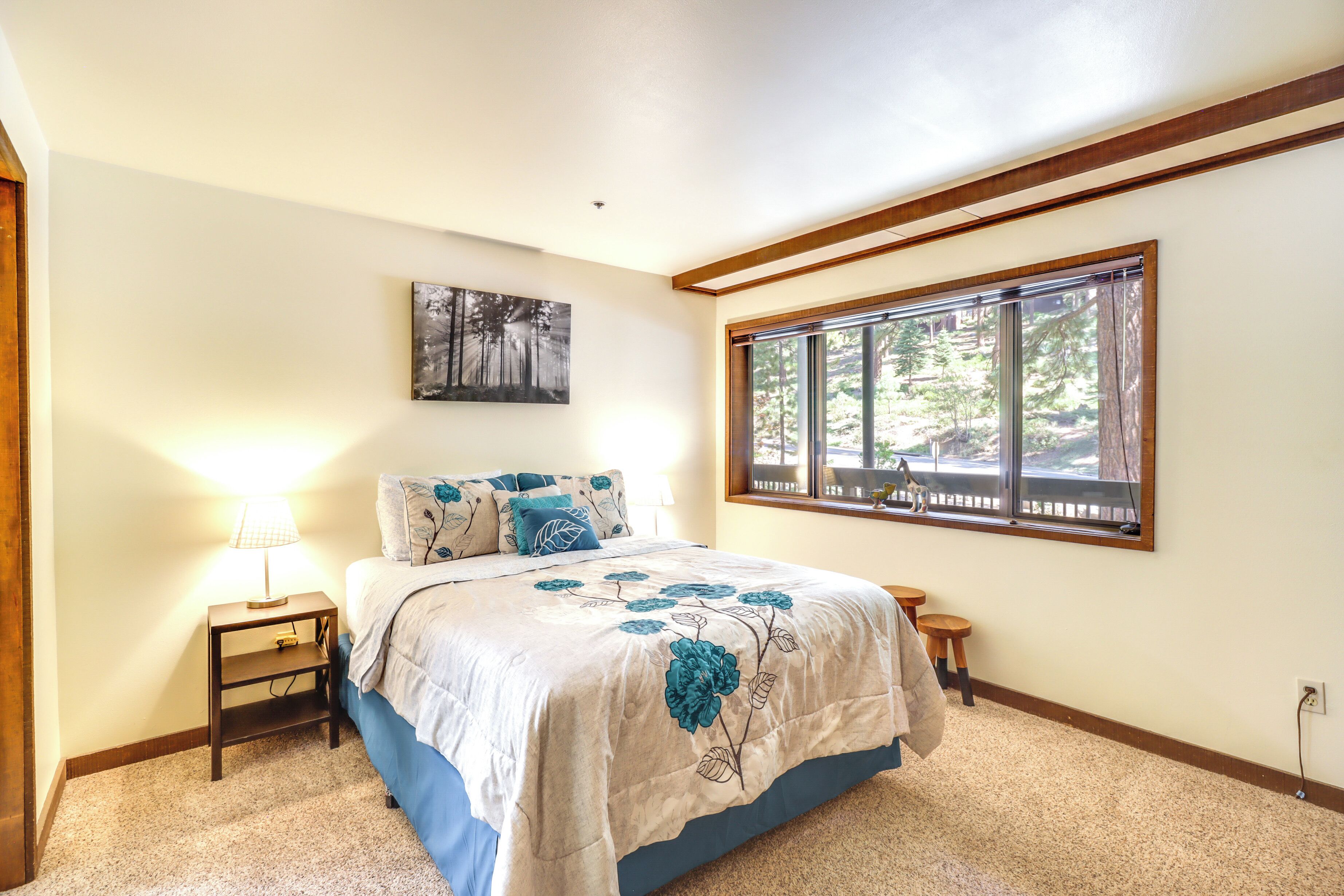 A bedroom with a large bed, a nightstand, a window with a view of trees, and a painting on the wall.