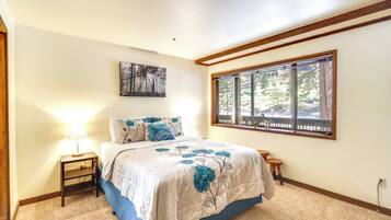 A bedroom with a large bed, a nightstand, a window with a view of trees, and a painting on the wall.