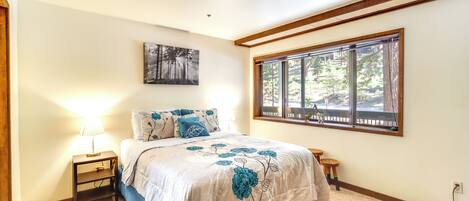 A bedroom with a large bed, a nightstand, a window with a view of trees, and a painting on the wall.