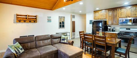 A cozy living room with a brown leather sectional sofa, wooden dining table, and chairs. A kitchen with wooden cabinets and modern appliances is visible in the background.