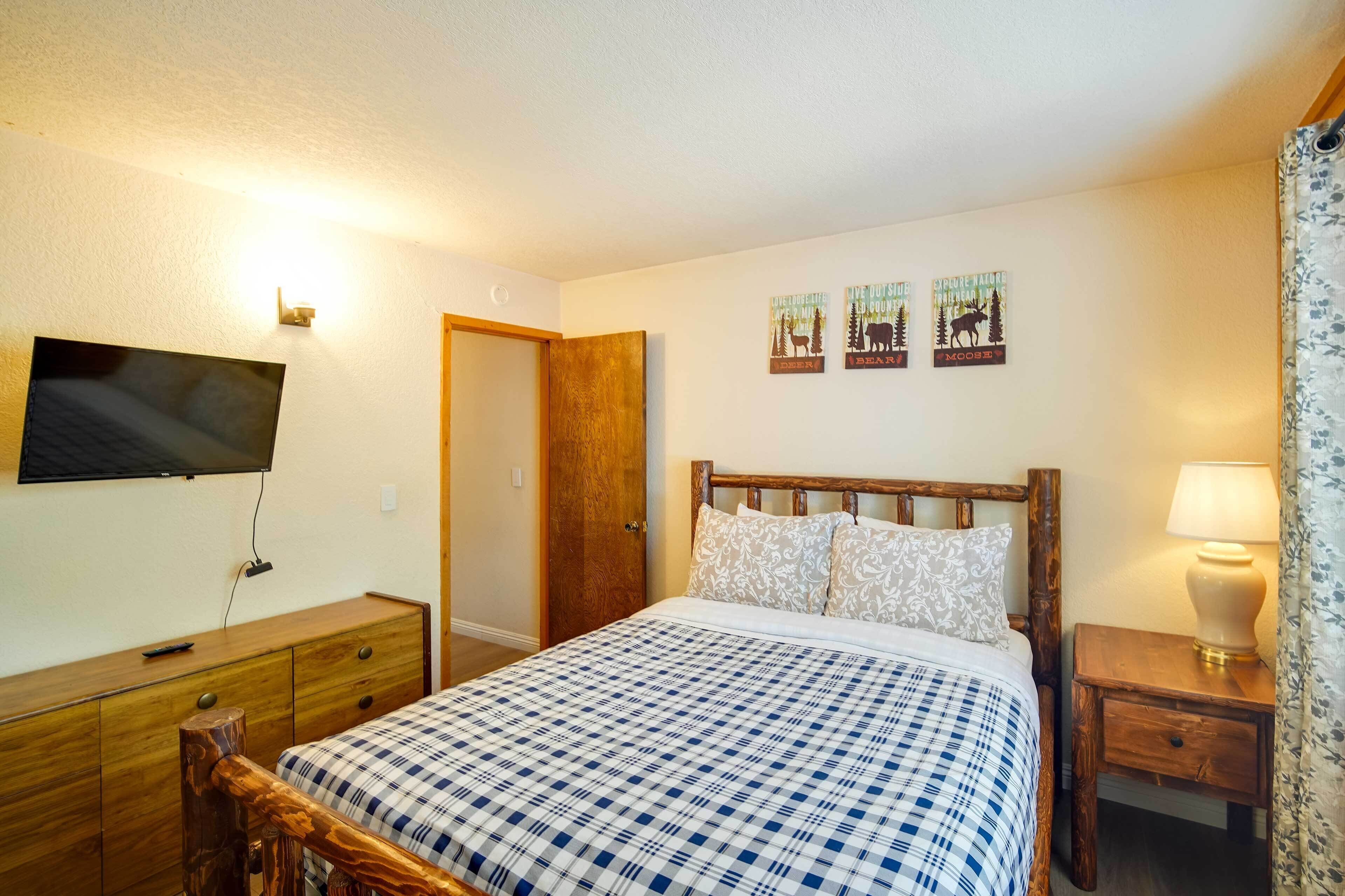 A bedroom with a wooden bed, a TV mounted on the wall, a wooden dresser, and a door leading to another room.