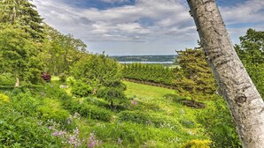 En frodig trädgård med ett stort utbud av växter och blommor, ett högt träd och en avlägsen utsikt över en vattenyta under en delvis molnig himmel.