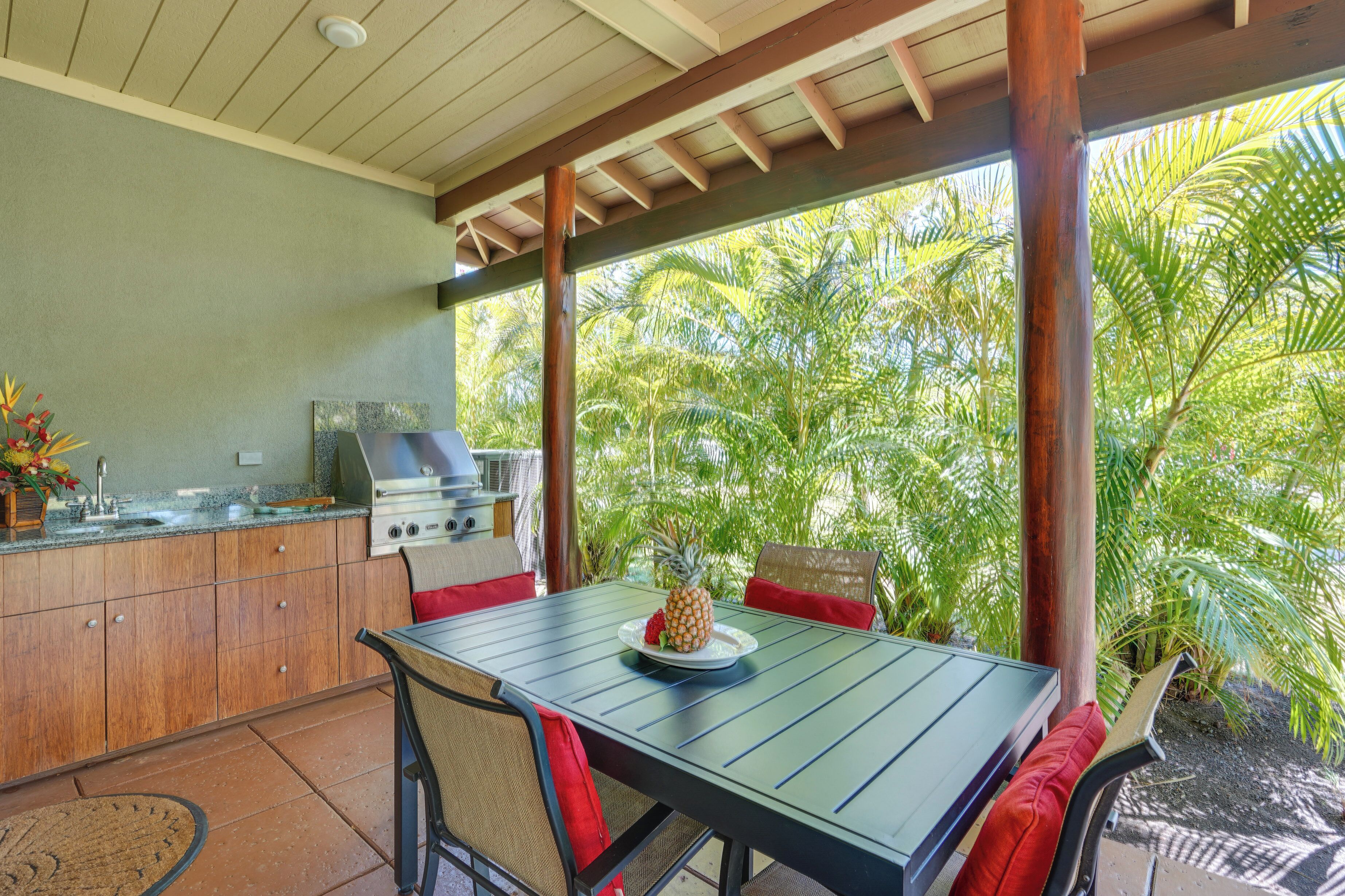 An outdoor kitchen with a grill, dining table, and chairs, surrounded by lush greenery.