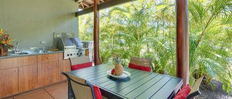 An outdoor kitchen with a grill, dining table, and chairs, surrounded by lush greenery.