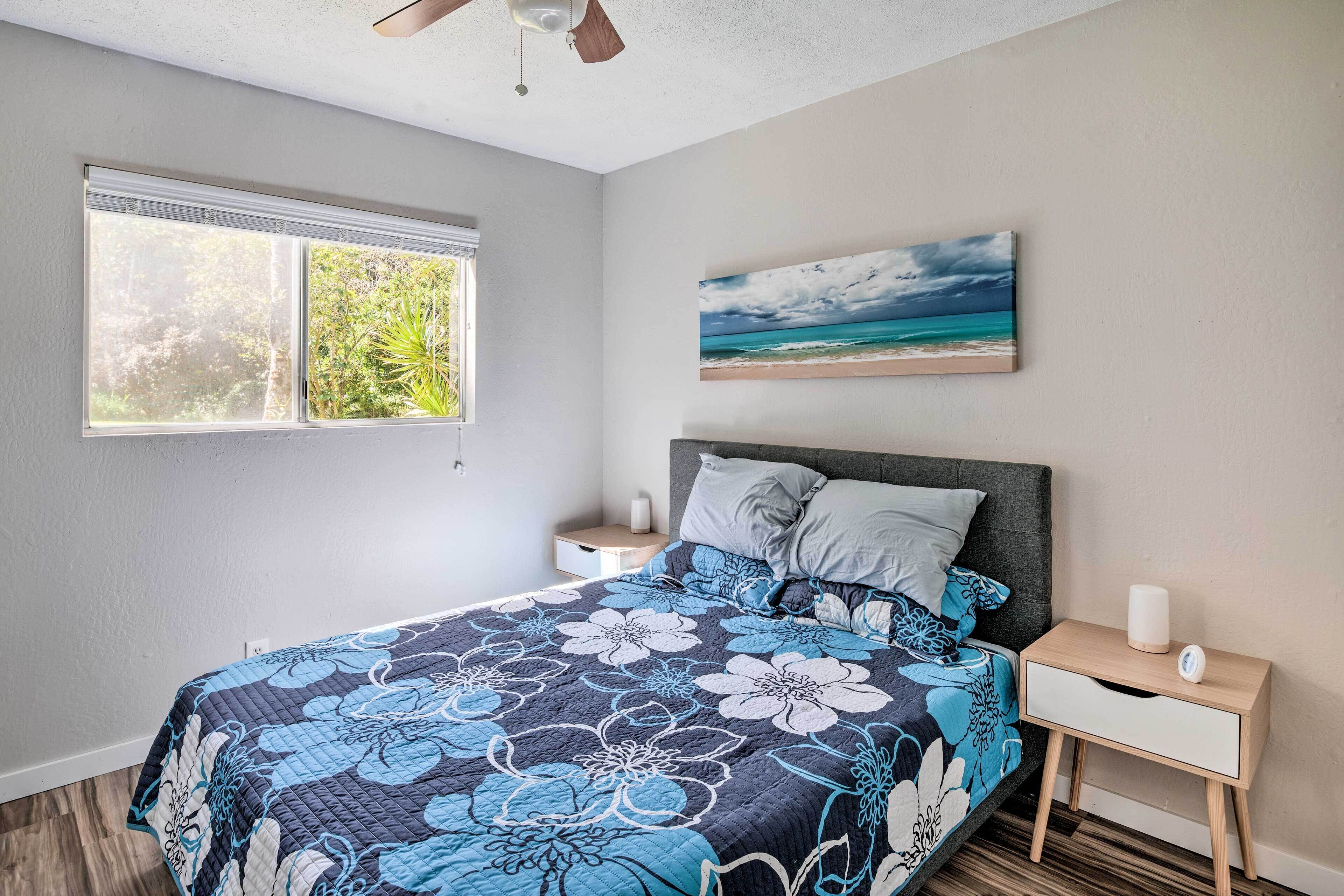 A bedroom with a large bed, a nightstand, a window with blinds, and a painting of a beach scene.