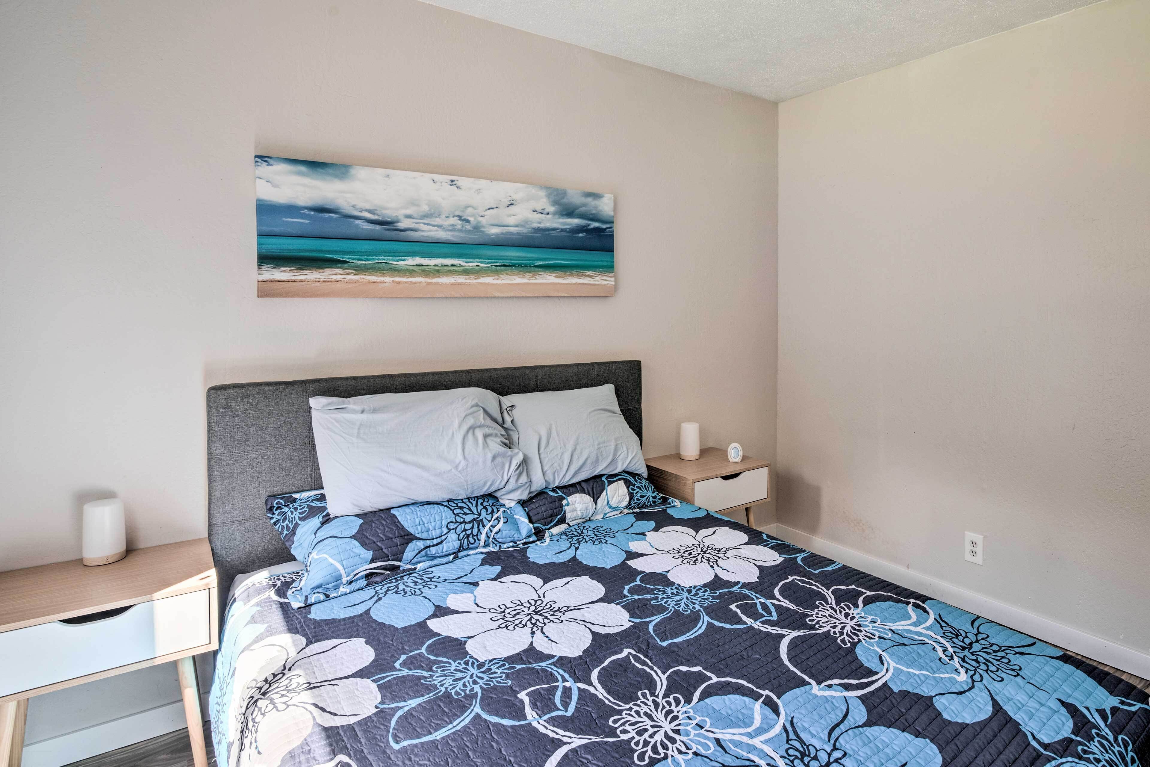 A bedroom with a bed, a nightstand, a bedside table, and a painting of a beach scene.