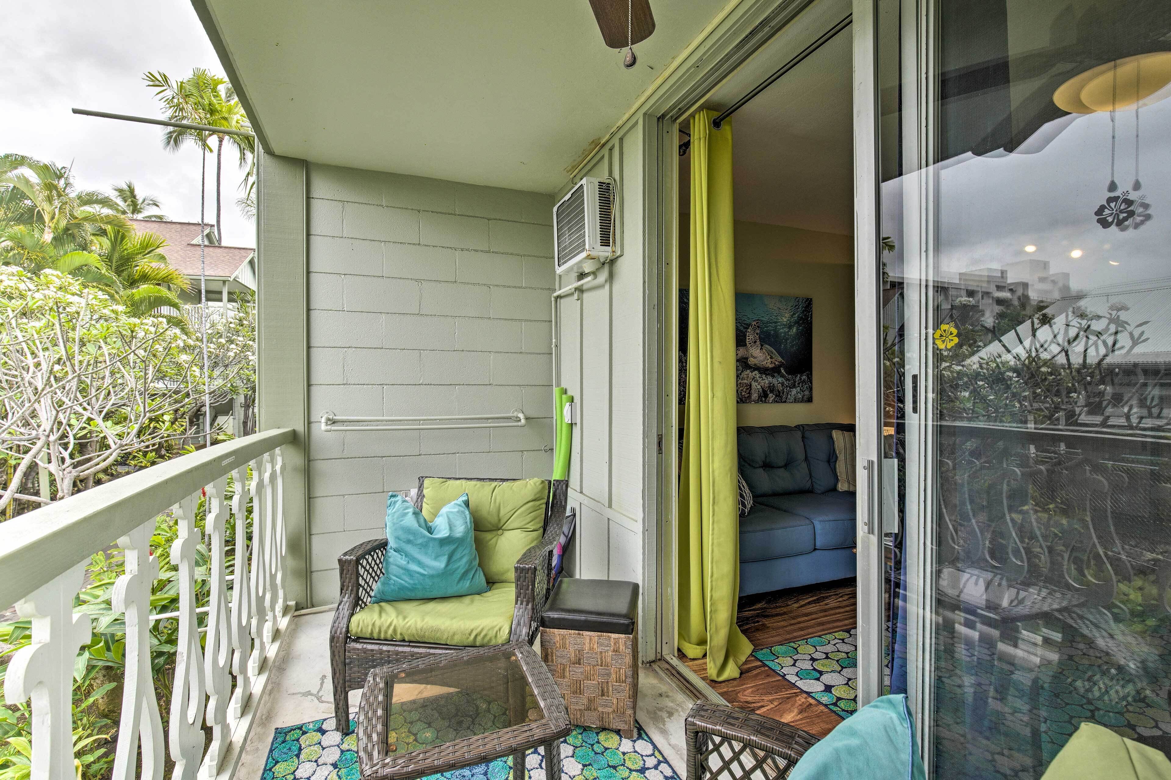 A balcony with a wicker chair, a small table, and a sliding glass door leading to a living room with a sofa and a painting.
