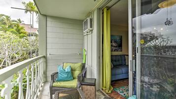 A balcony with a wicker chair, a small table, and a sliding glass door leading to a living room with a sofa and a painting.