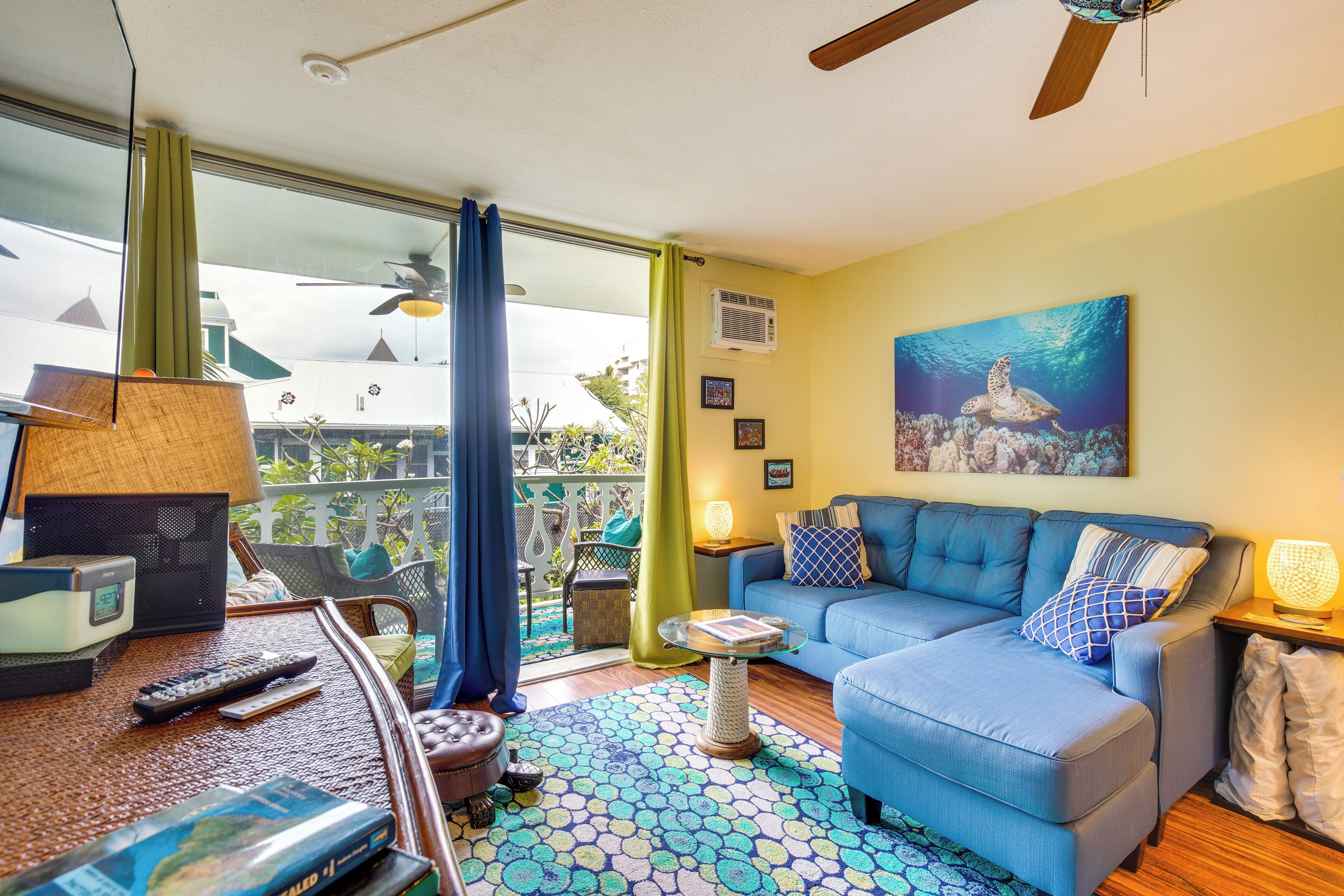 A living room with a blue sofa, a coffee table, and a ceiling fan. There is a window with curtains, a painting on the wall, and a lamp on a side table.