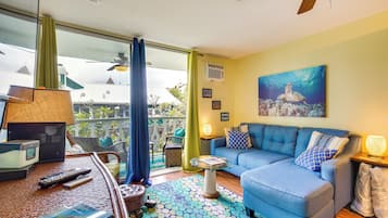 A living room with a blue sofa, a coffee table, and a ceiling fan. There is a window with curtains, a painting on the wall, and a lamp on a side table.