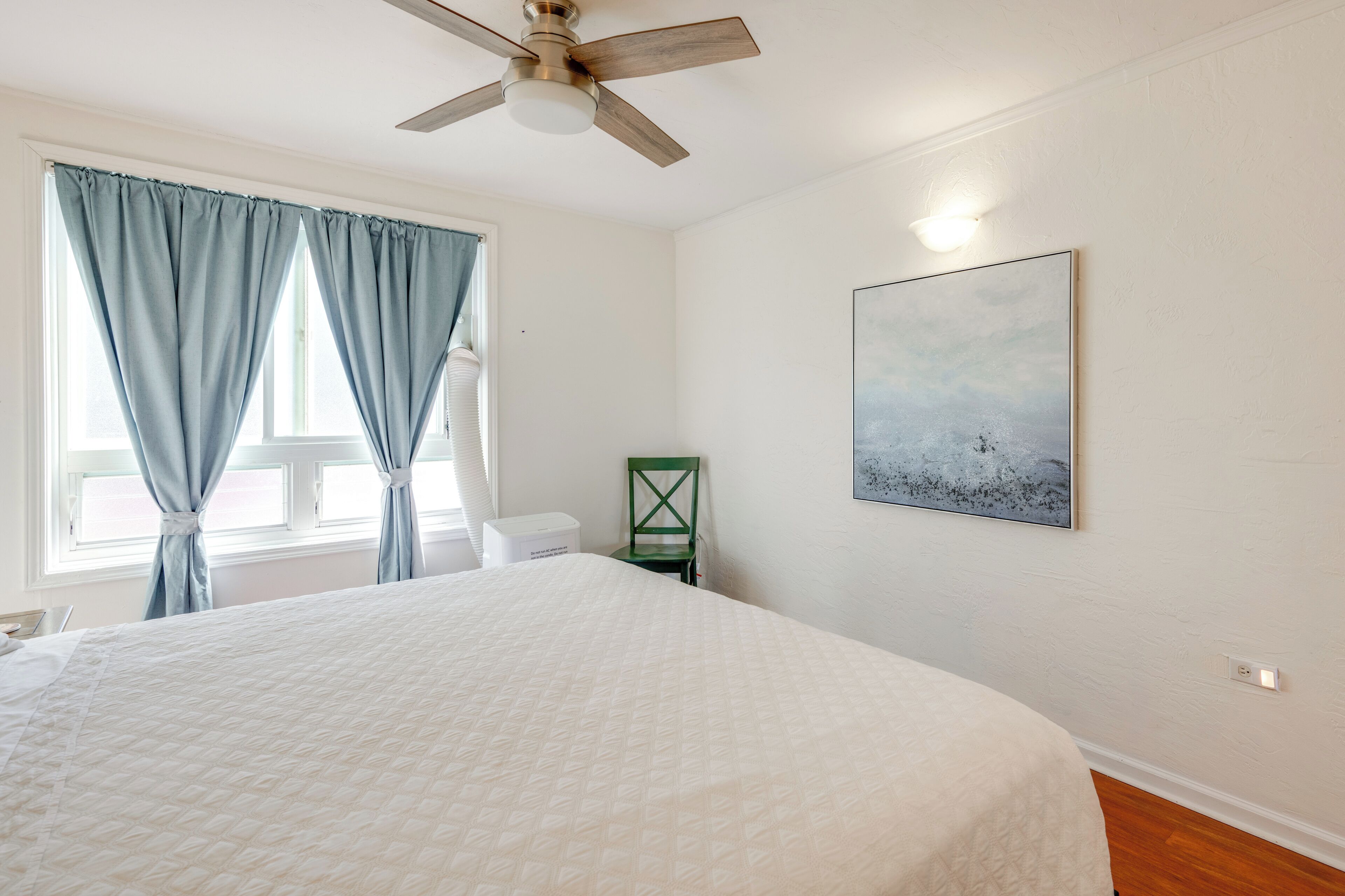 Quarto com cama, ventoinha no teto, janela com cortinas, cadeira e uma obra de arte na parede.