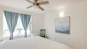 Une chambre à coucher comprenant un lit, un ventilateur de plafond, une fenêtre avec des rideaux, une chaise et une œuvre d’art accrochée au mur.