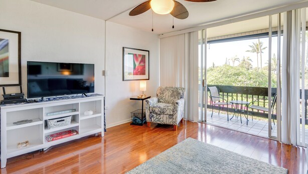 A living room with a flat-screen TV, a ceiling fan, a chair, and a view of a balcony with outdoor furniture.