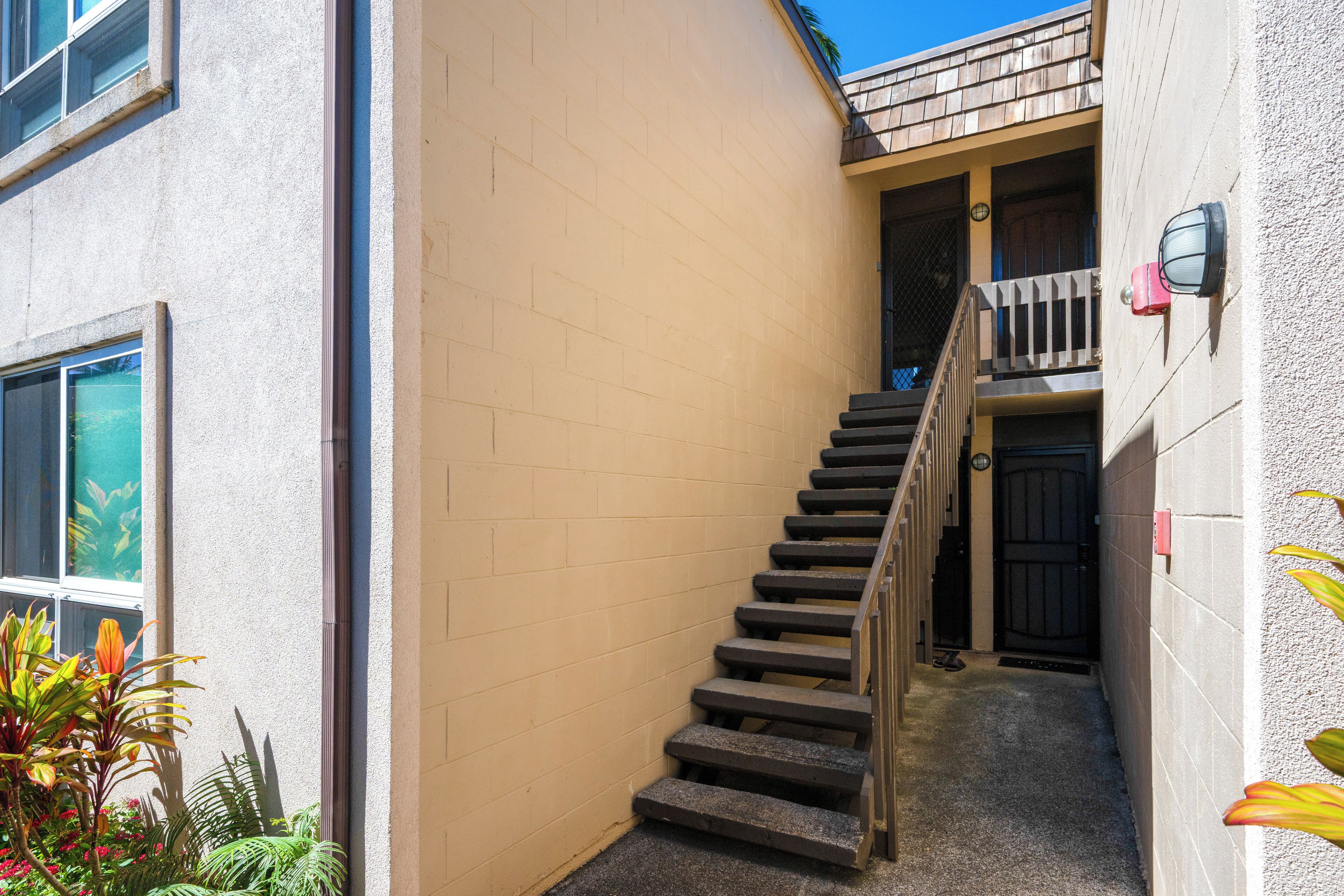 A narrow stairway leading to a balcony with a wooden railing, a door, and a small outdoor area with plants.