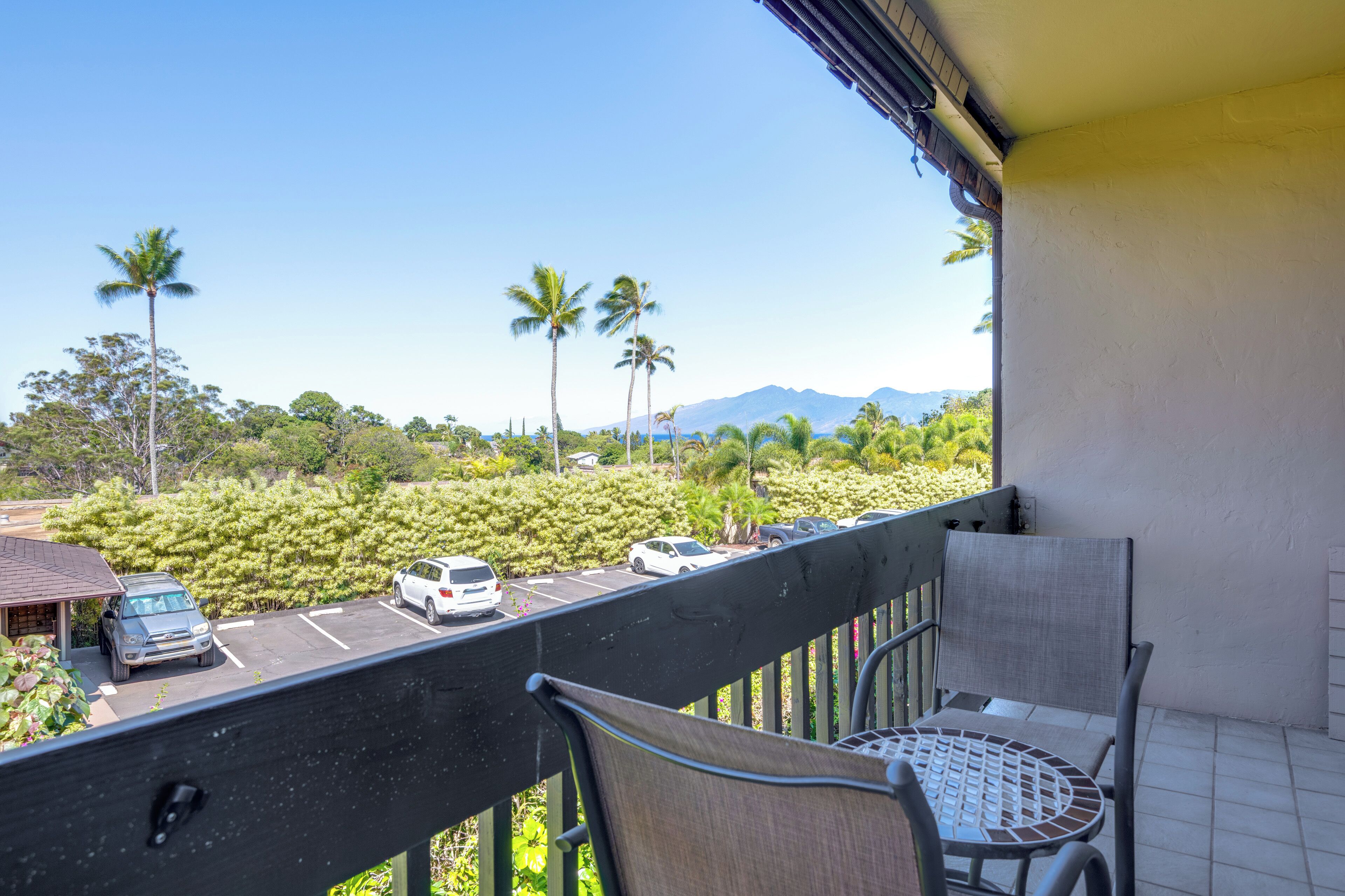 A balcony with a table and chairs, overlooking a parking lot with cars and palm trees, with mountains in the background.
