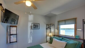 Ett sovrum med en säng, en takfläkt, ett fönster med utsikt över vattnet, en väggmonterad TV och en hylla med handdukar.