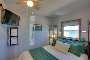 A bedroom with a bed, a ceiling fan, a window with a view of the water, a wall-mounted TV, and a shelf with towels.