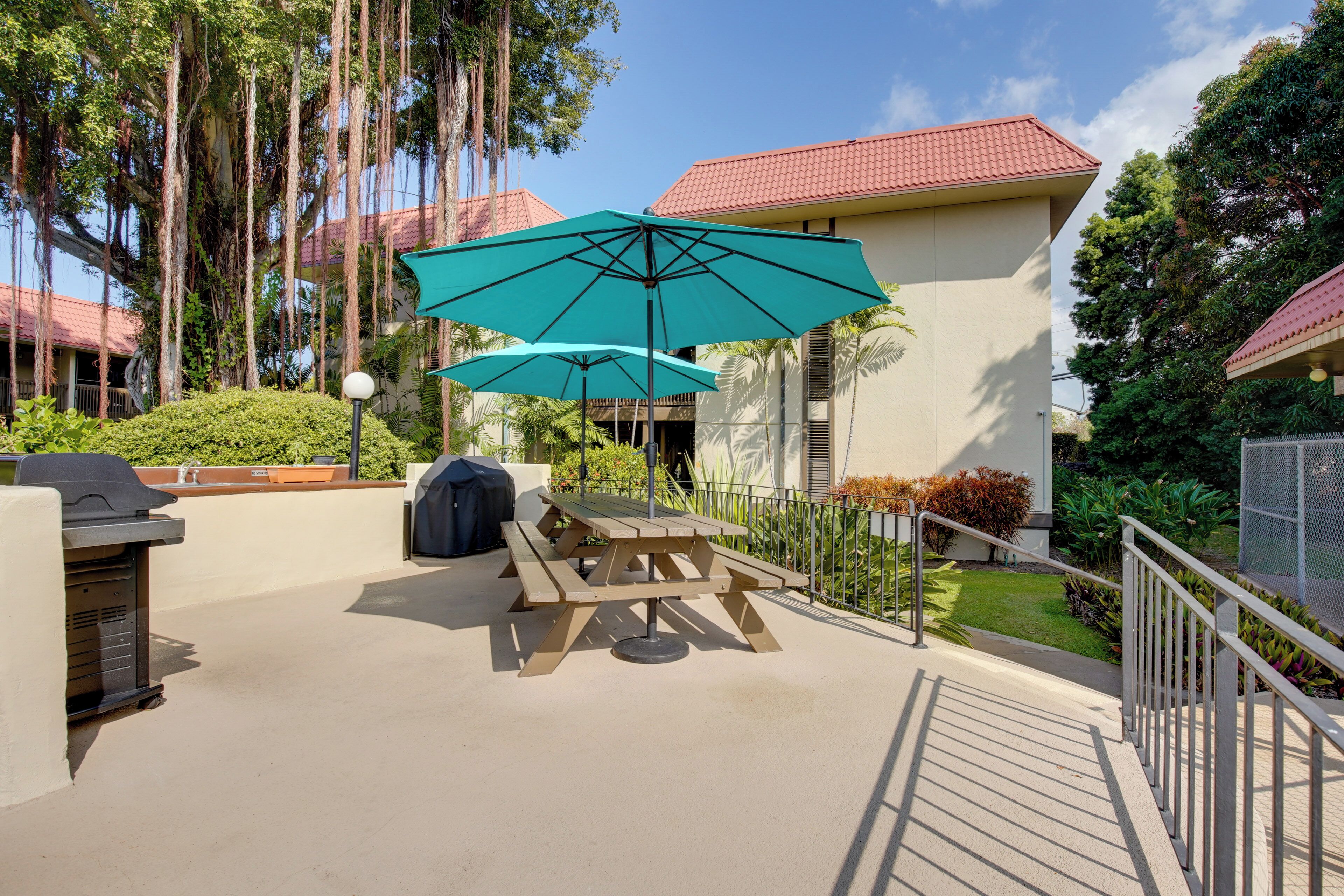An outdoor patio with a picnic table and a large umbrella, surrounded by greenery and a building with a red roof.