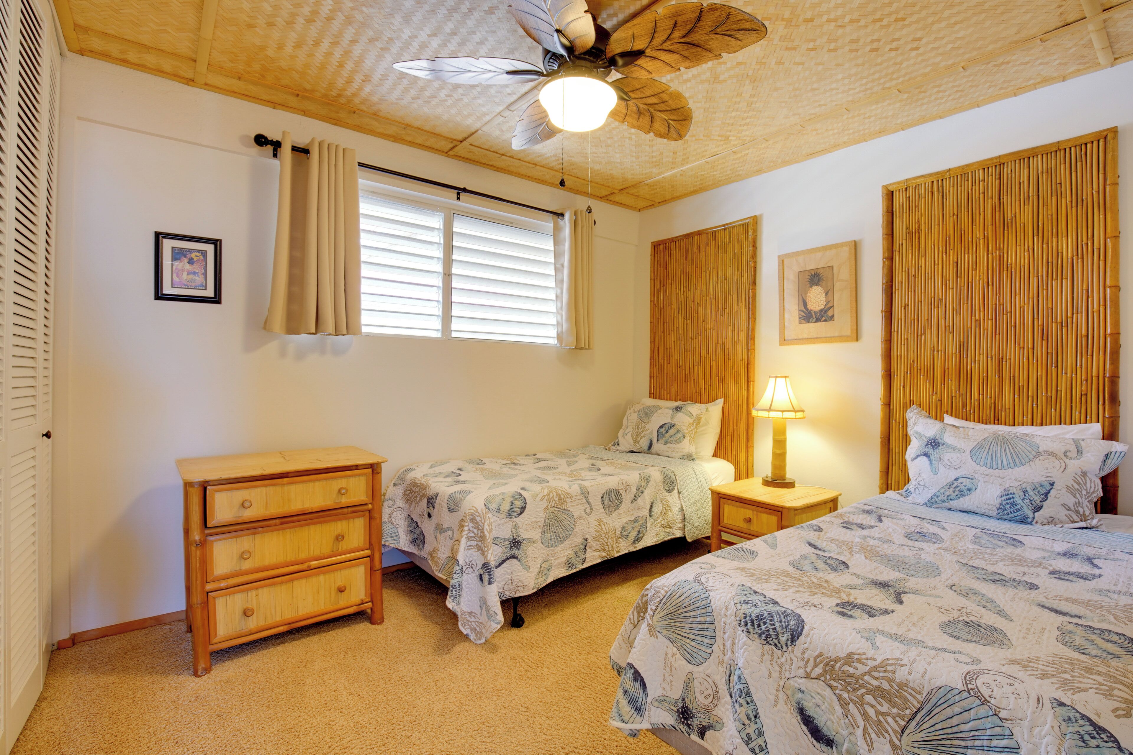 Un dormitorio con dos camas, un ventilador de techo, una ventana con cortinas, una lámpara de noche y un aparador de madera.