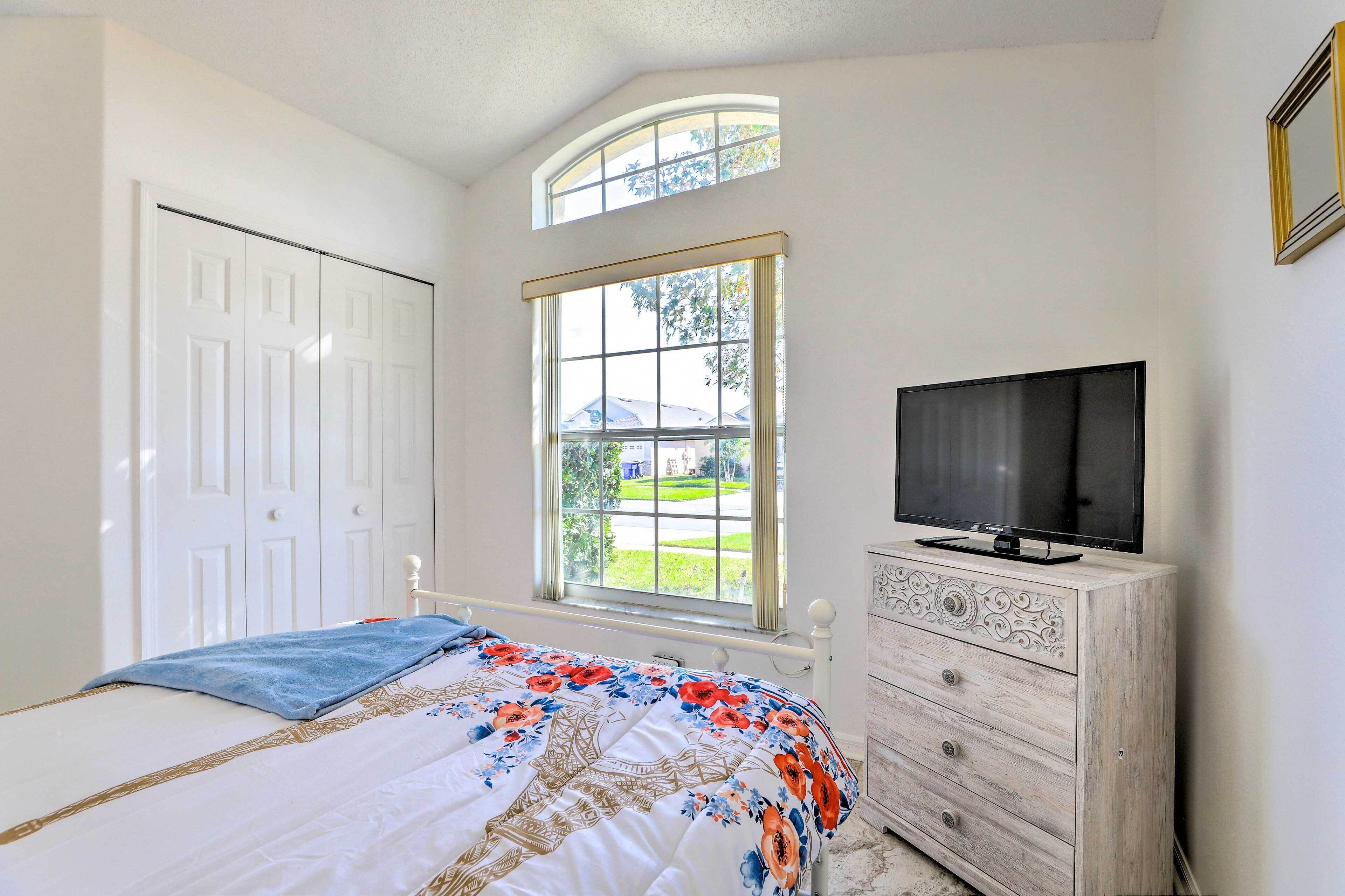 A bedroom with a bed, a dresser, a television, and a window with a view of a house and lawn.