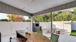 Un patio exterior con mesa y sillas de madera, chimenea de piedra y valla blanca.