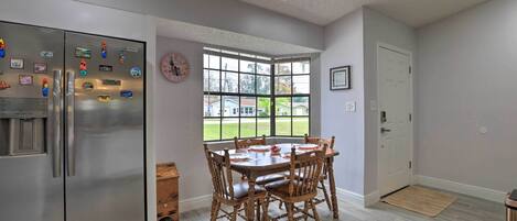 A kitchen with a stainless steel refrigerator, wooden dining table with chairs, a window with a view of houses, and a white door.