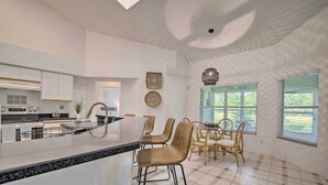 Een moderne keuken met een groot eiland, een eethoek met rieten stoelen en grote ramen met zonwering.