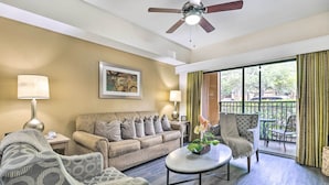 Una sala de estar con un sofá, sillones, una mesa de centro y un ventilador de techo. Hay una ventana con cortinas y vistas al exterior.