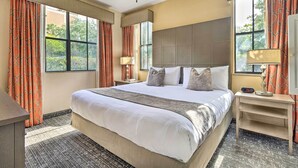 Habitación de hotel con una cama grande, un escritorio, una mesita de noche, un ventilador de techo y ventanas amplias con cortinas.
