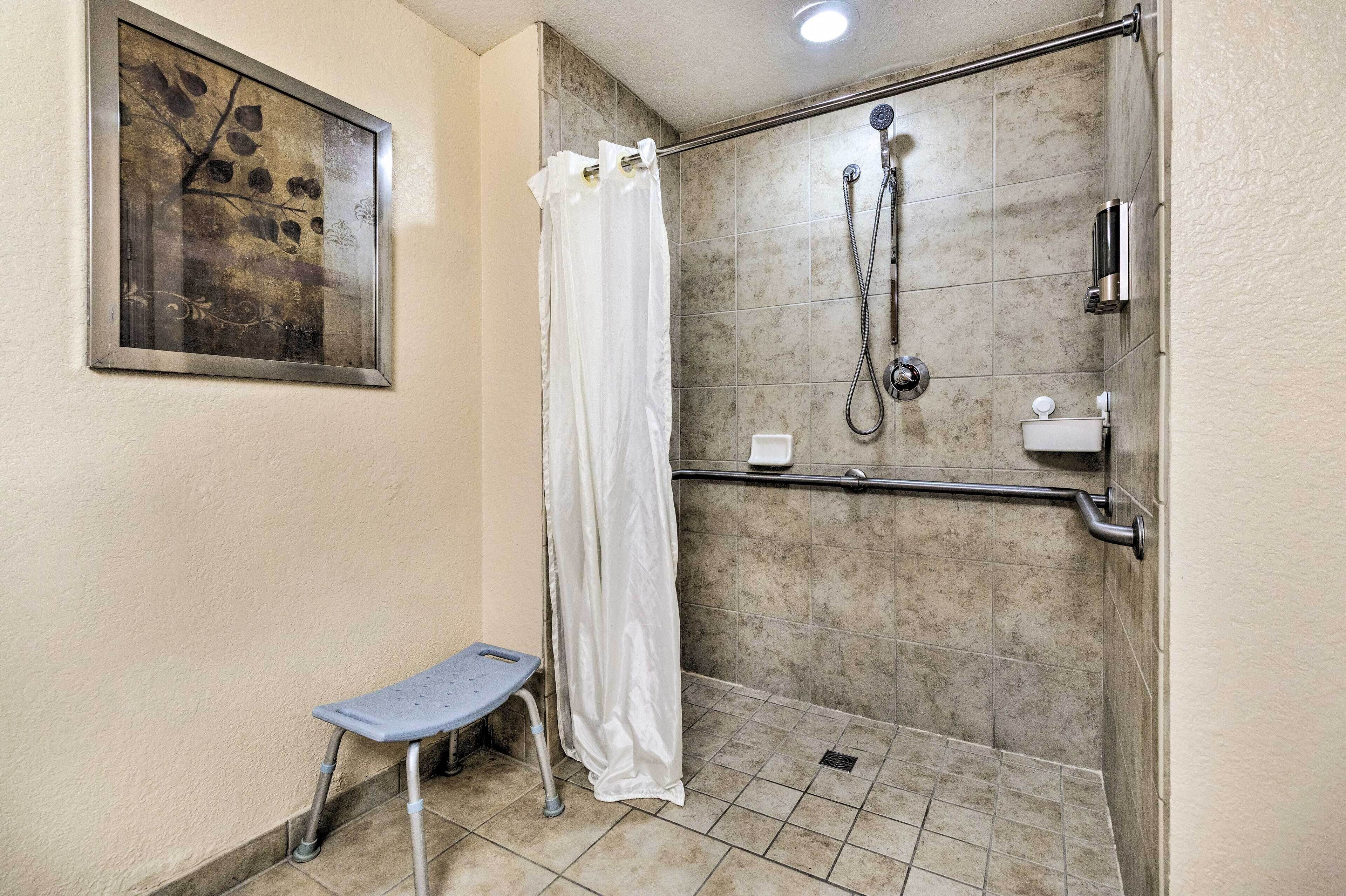 A shower area with a glass door, a white shower curtain, a handheld showerhead, a handheld shower attachment, a grab bar, and a small step stool.