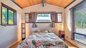 Een slaapkamer met een houten plafond, een bed met een bloemenbedekking, twee ramen met gordijnen, en een nachtkastje met een lamp.